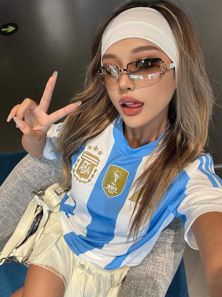 Foto de mulher asiática usando camisa da Argentina, óculos de sol, faixa bege no cabelo e alongando o pescoço 