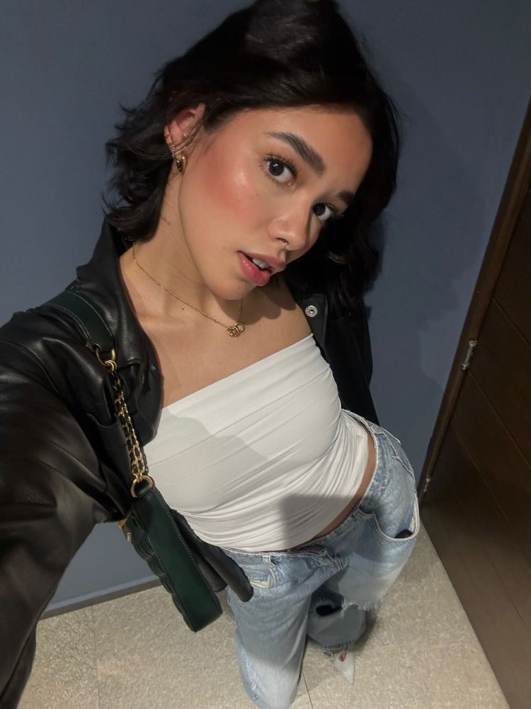 Selfie de mulher de pele clara com jaqueta preta, top branco e calça jeans, tirada de cima 