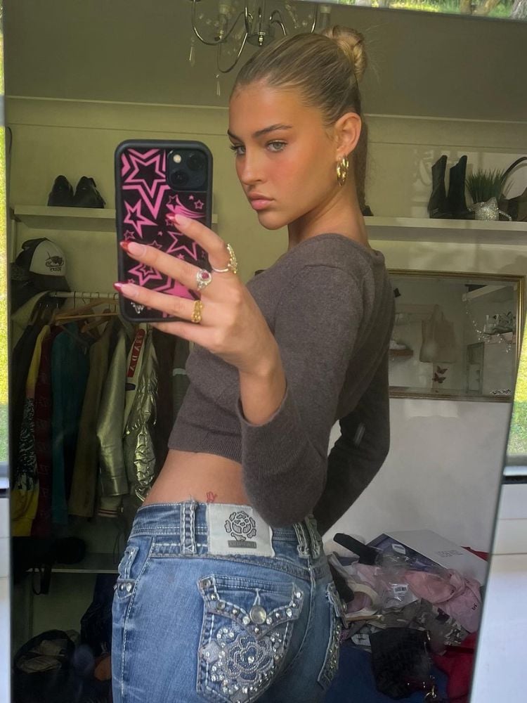 Mulher de pele clara usando cropped cinza e calça jeans tirando selfie de costas para o espelho 