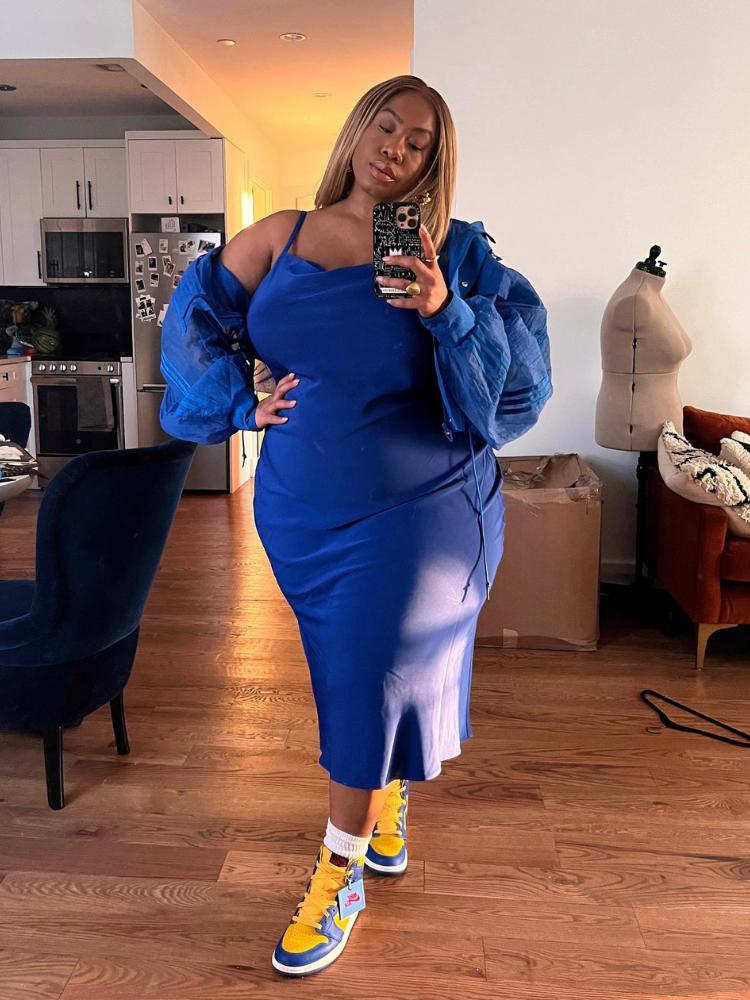 Mulher de pele negra usando vestido midi azul e casaco também azul tirando foto no espelho com as pernas cruzadas 