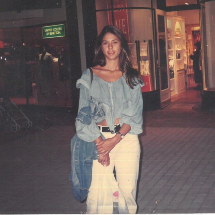 Foto antiga de Priscila Belfort usando calça branca e blusa jeans, em frente a uma loja 