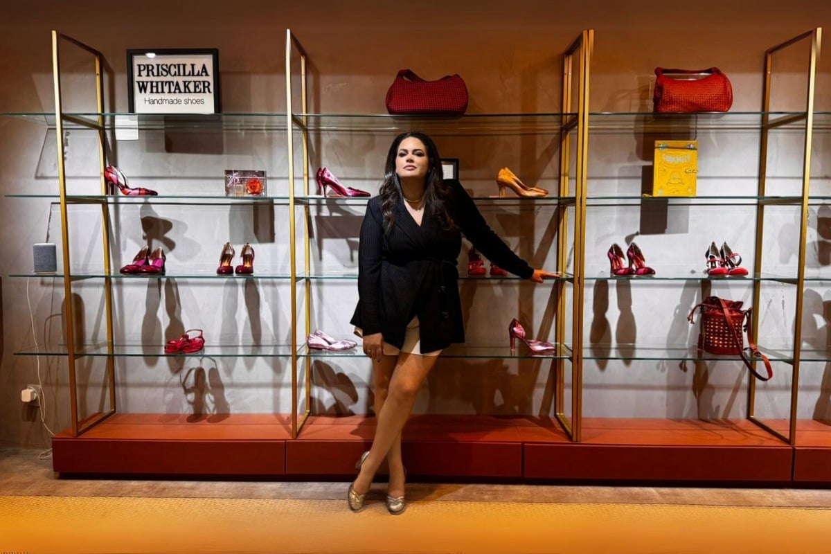 Priscilla Whitaker usando blazer preto fechado e shorts bege. Ela está em frente a prateleiras de sua loja, com sapatos artesanais expostos