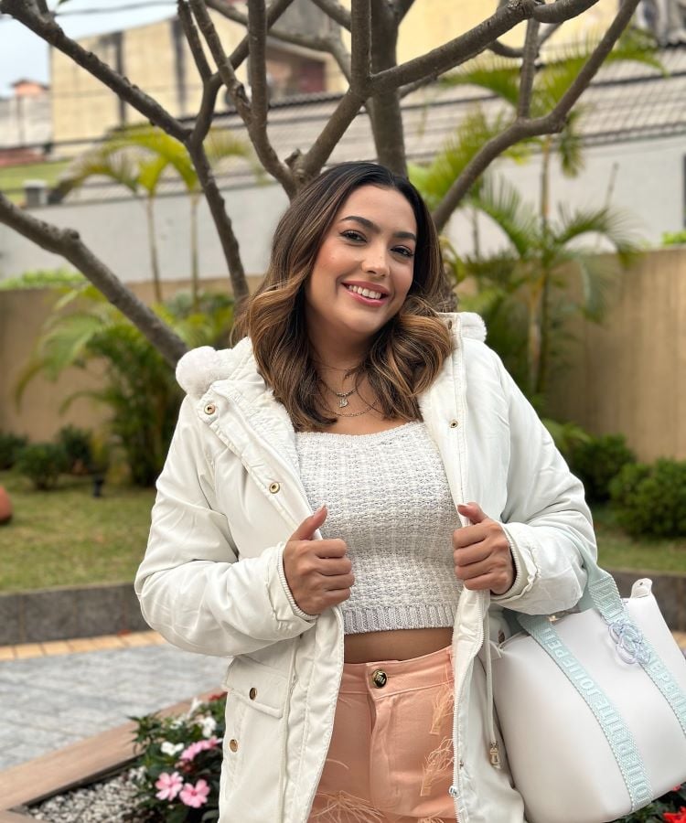 A imagem mostra uma mulher sorridente em um jardim. Ela veste uma jaqueta branca, um top cinza e calças de cor clara. Ao fundo, há árvores e plantas. Recentemente, procedimentos estéticos têm sido um tema de interesse em programas como A Fazenda 16.