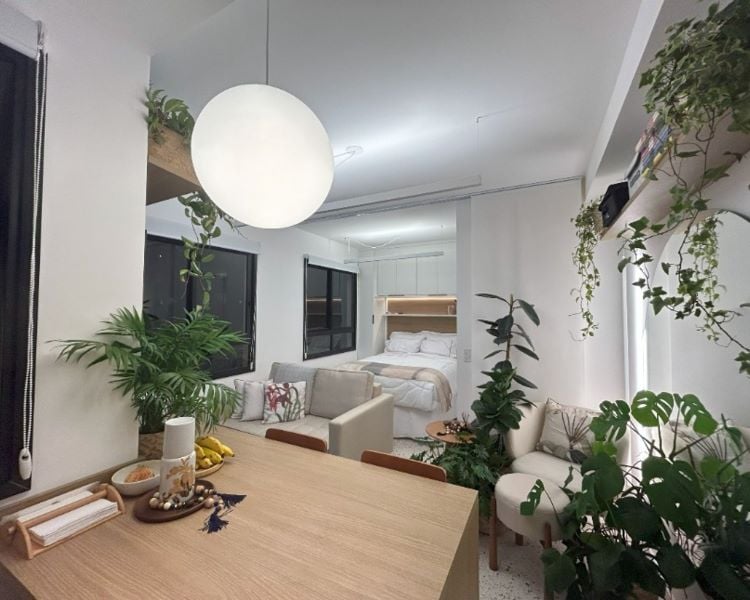 A imagem mostra um apartamento compacto, integrado e bem iluminado. O espaço combina sala de estar, jantar e quarto. Uma mesa de madeira clara com frutas e objetos decorativos está em primeiro plano, com cadeiras ao redor. Ao fundo, há um sofá pequeno e uma cama arrumada com roupas de cama brancas. Plantas decorativas estão espalhadas pelo ambiente, tanto no chão quanto em prateleiras, dando um toque natural ao espaço. Grandes janelas com moldura preta permitem a entrada de luz natural. Uma luminária pendente esférica ilumina o ambiente de forma suave, criando uma atmosfera aconchegante e moderna.