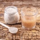 A imagem mostra psyllium em três formas: um pote de vidro cheio de psyllium em pó, uma colher de madeira contendo o mesmo pó, e um copo com água onde o psyllium foi misturado, formando uma substância gelatinosa. Todos os itens estão sobre uma superfície de madeira rústica, destacando a textura do psyllium.