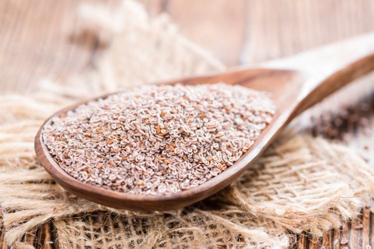 A imagem mostra uma colher de madeira cheia de sementes de psyllium, dispostas sobre uma superfície de tecido rústico. As sementes são pequenas, de cor marrom claro, e possuem uma textura fina e granulada. O fundo desfocado destaca a simplicidade e naturalidade dos ingredientes, sugerindo um ambiente de cozinha ou preparo de alimentos saudáveis.