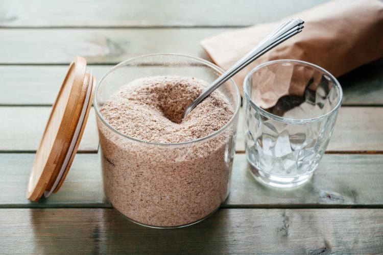 A imagem apresenta um pote de vidro aberto, cheio de psyllium em pó, com uma colher de metal mergulhada nele. Ao lado, há um copo de vidro vazio e uma embalagem de papel pardo. Tudo está disposto sobre uma superfície de madeira em tom verde acinzentado, criando um ambiente simples e natural.