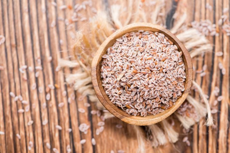 A imagem mostra um pequeno recipiente de madeira cheio de sementes de psyllium, colocado sobre um fundo de madeira rústica com algumas fibras de juta. As sementes são pequenas e de cor clara, com uma textura levemente granulada. Algumas sementes estão espalhadas ao redor, criando um aspecto natural e orgânico.