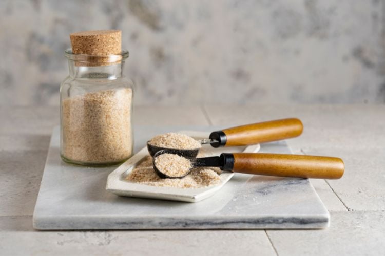 A imagem mostra um pote de vidro com tampa de cortiça cheio de psyllium em pó. Ao lado, há uma bandeja retangular de cerâmica com duas colheres de cabo de madeira, também cheias de psyllium. Tudo está disposto sobre uma superfície de mármore clara, com um fundo de parede texturizada em tons neutros.