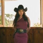 Gizelly Bicalho usando vestido vinho de um ombro só, cinto preto com ferragens em ouro velho e chapéu de cowboy marrom. O cenário da foto se assemelha a um celeiro