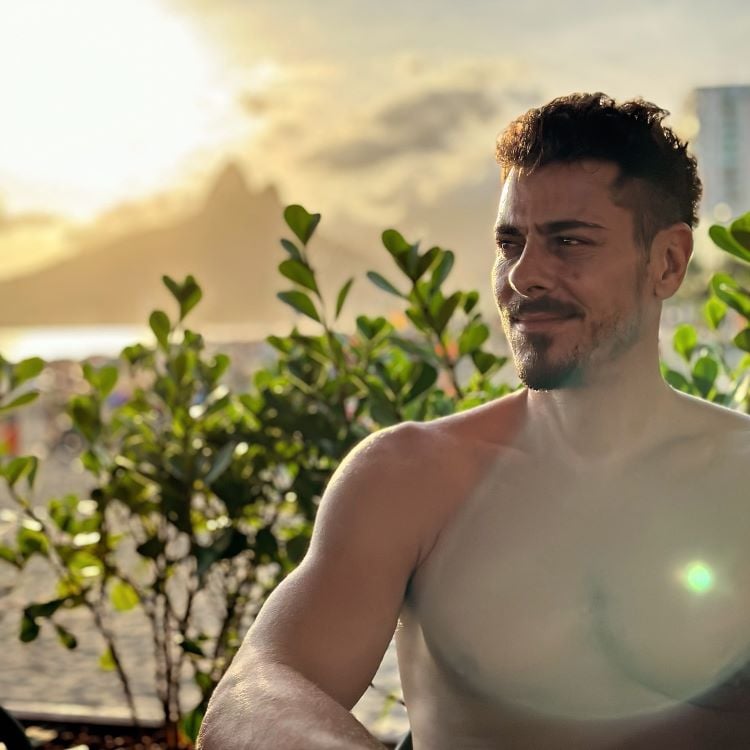 A imagem mostra Sacha Bali sem camisa, sentado em um ambiente ao ar livre. Ao fundo, há vegetação e o sol se pondo, criando uma iluminação dourada. Montanhas são visíveis à distância, sugerindo uma paisagem costeira. O homem está olhando para o lado, com uma expressão tranquila.