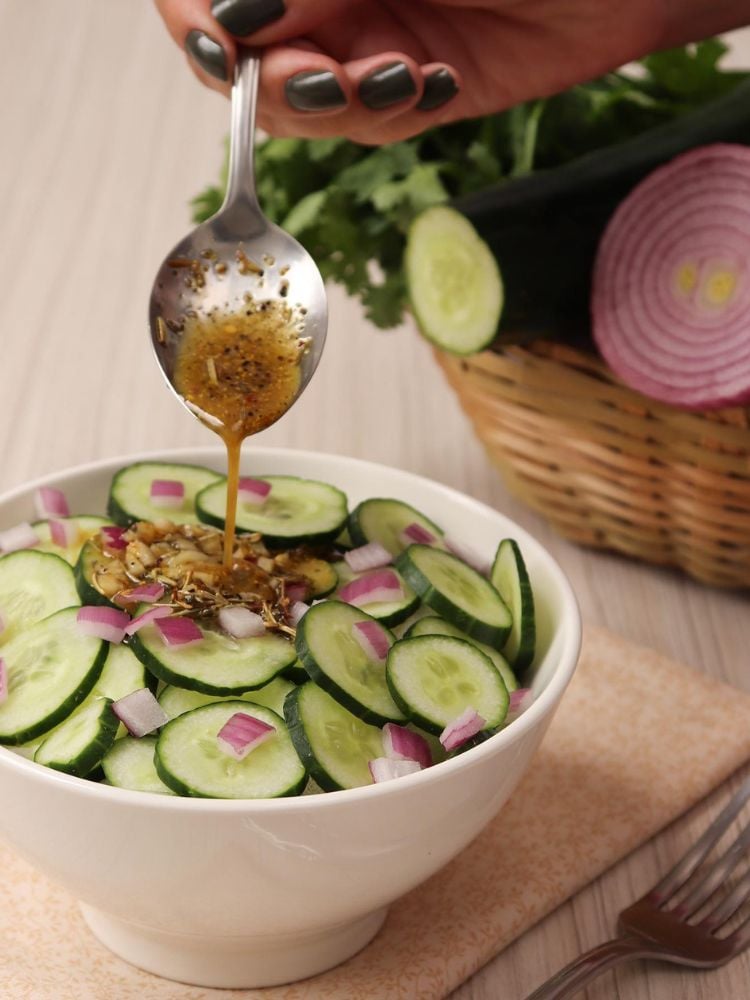 Pessoa de pele clara colocando molho marrom em salada de pepino com cebola roxa em bowl branco 