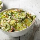 Bowl branco com salada de pepino, molho cremoso e temperos sobre mesa e ao lado de garfo