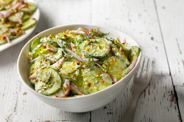 Bowl branco com salada de pepino, molho cremoso e temperos sobre mesa e ao lado de garfo