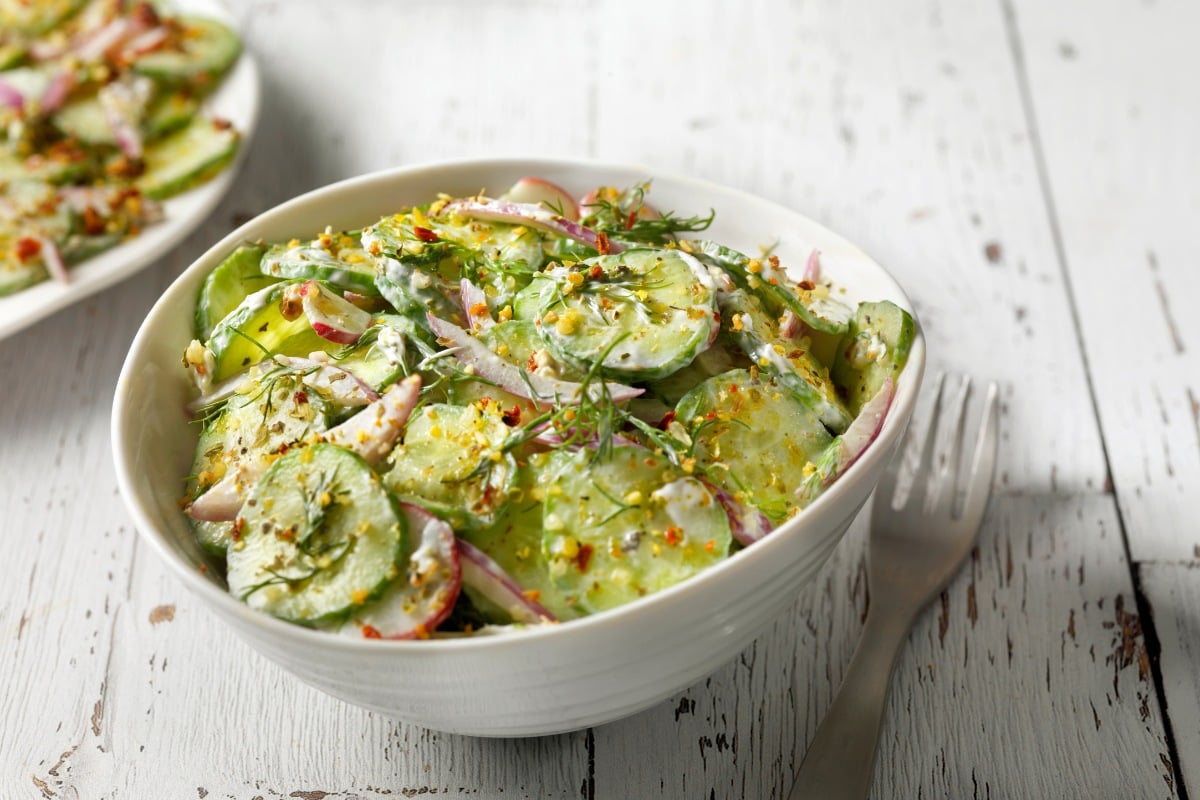 Bowl branco com salada de pepino, molho cremoso e temperos sobre mesa e ao lado de garfo