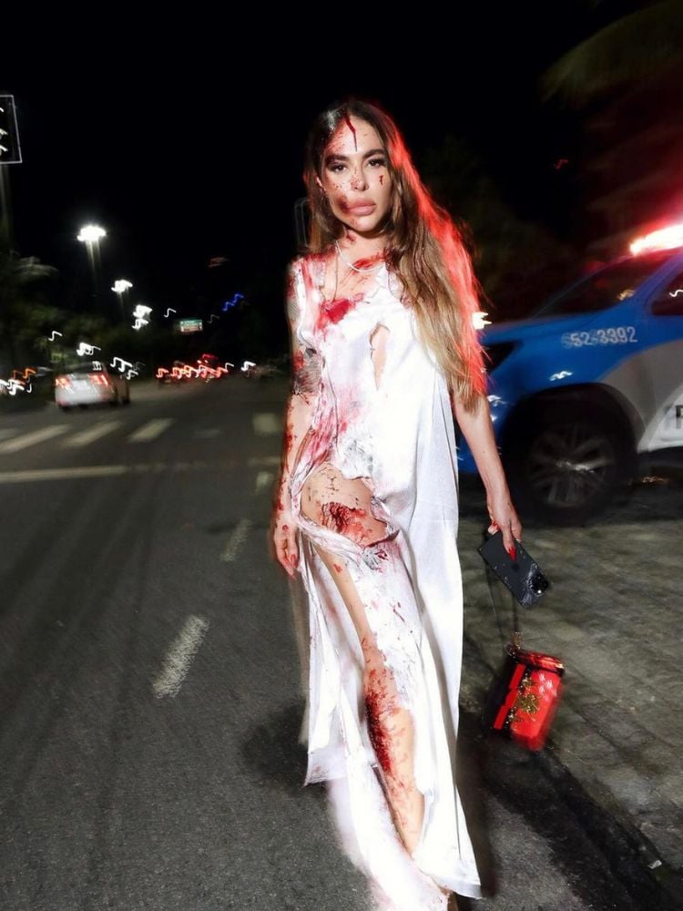 Mulher de pele clara andando na rua, usando maquiagem de Halloween com roupa branca rasgada e corpo com sangue falso 