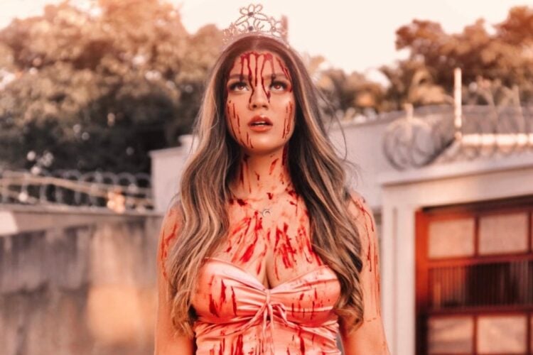 Jovem de pele clara com vestido rosado, coroa e sangue falso por todo o corpo