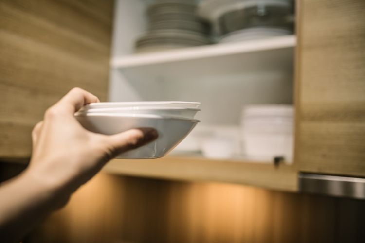 A imagem mostra uma mão segurando um conjunto de pratos brancos, prestes a colocá-los em um armário de cozinha. O armário é de madeira clara, e no interior dele há mais pratos e tigelas organizados. A iluminação suave cria um ambiente acolhedor e bem cuidado.