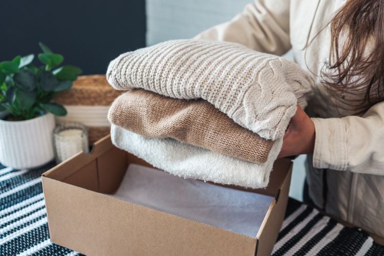 A imagem mostra uma pessoa colocando roupas dobradas em uma caixa de papelão. As roupas são de cores neutras, como bege e branco, e parecem ser peças de inverno, como suéteres. Ao lado, há uma planta em um vaso branco e uma superfície coberta por um tecido listrado em preto e branco. A cena transmite uma sensação de organização e cuidado.