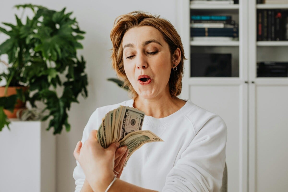 A mulher está em um ambiente doméstico, segurando um maço de dinheiro com expressão de surpresa e satisfação. Ela veste uma blusa branca. Ao fundo, há uma planta verde e uma estante com livros, sugerindo prosperidade e conforto.