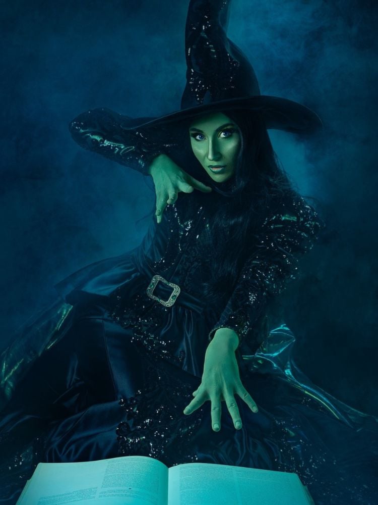 Pessoa fantasiada de bruxa Elphaba, com pele pintada de verde, vestido preto com brilho e chapéu de bruxa 