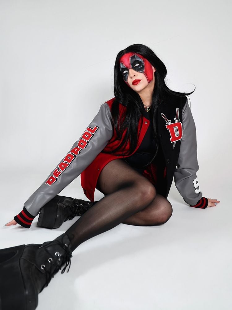 Mulher de pele clara sentada no chão, usando jaqueta do Deadpool, roupa preta, coturno e maquiagem que imita a máscara do herói 