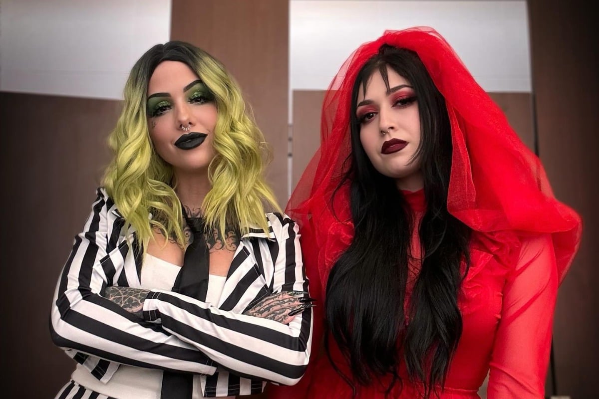 Duas mulheres fantasiadas, sendo uma delas de cabelo esverdeado e terno listado preto e branco como de Beetlejuice e outra mulher com vestido e véu vermelho, fantasiada da personagem Lydia