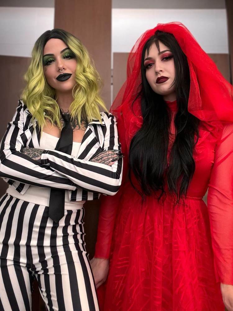 Duas mulheres fantasiadas, sendo uma delas de cabelo esverdeado e terno listado preto e branco como de Beetlejuice e outra mulher com vestido e véu vermelho, fantasiada da personagem Lydia 