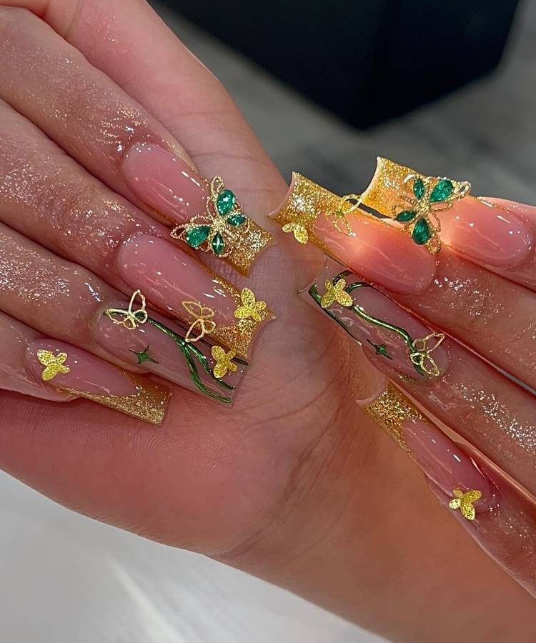 A imagem mostra mãos com unhas longas e quadradas, decoradas com um esmalte transparente. As unhas têm detalhes dourados brilhantes e pequenos desenhos de borboletas em verde e dourado. As borboletas estão distribuídas artisticamente, criando um efeito luxuoso. A pele das mãos brilha, sugerindo o uso de um produto cintilante, destacando ainda mais o design das unhas.