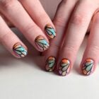 A imagem mostra mãos com unhas decoradas em um estilo que lembra vitrais. As unhas têm desenhos coloridos em tons de azul, laranja e rosa, com contornos pretos, criando um efeito de mosaico. As mãos estão sobre uma superfície clara, e uma delas usa um anel dourado, adicionando um toque de elegância.