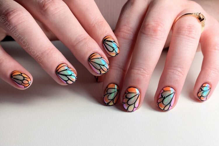 A imagem mostra mãos com unhas decoradas em um estilo que lembra vitrais. As unhas têm desenhos coloridos em tons de azul, laranja e rosa, com contornos pretos, criando um efeito de mosaico. As mãos estão sobre uma superfície clara, e uma delas usa um anel dourado, adicionando um toque de elegância.