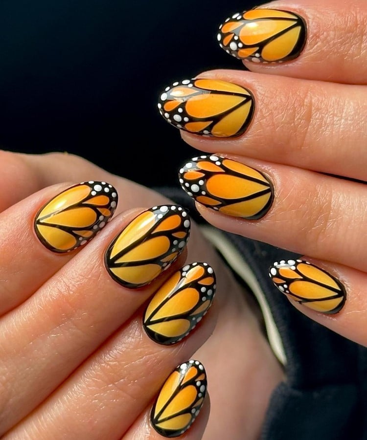 A imagem mostra unhas decoradas com um design inspirado em asas de borboleta. As unhas são pintadas em tons de laranja e amarelo, com contornos pretos que criam formas semelhantes a pétalas. Pequenos pontos brancos adornam as bordas, imitando detalhes delicados das asas. O visual é vibrante e artisticamente detalhado, destacando-se sobre um fundo escuro.