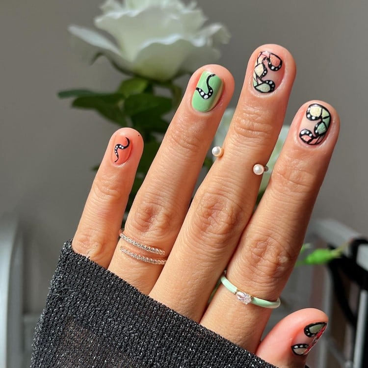 A imagem mostra uma mão com unhas curtas e arredondadas, decoradas com um design de borboletas em tons de verde e rosa. As unhas têm contornos pretos e detalhes em branco, criando um efeito de vitral. A mão usa anéis finos, um com pérolas. Ao fundo, há uma flor branca desfocada, destacando o visual artístico das unhas.