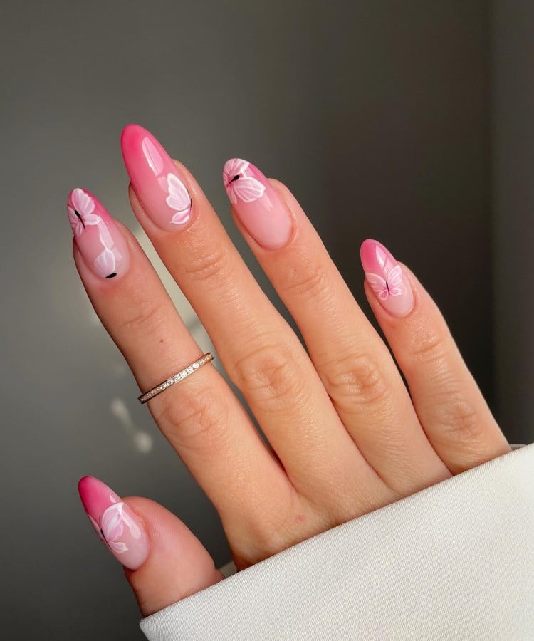 A imagem mostra uma mão com unhas longas e ovais, decoradas com um degradê de rosa claro para rosa escuro nas pontas. Cada unha tem um desenho de borboleta branca, acrescentando um toque delicado. A mão está adornada com um anel fino e dourado no dedo médio. O fundo é suave, destacando as unhas.