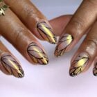 A imagem mostra mãos com unhas decoradas em formato de asas de borboleta. As unhas têm um design em tons de dourado e lilás, com contornos pretos e detalhes em branco nas pontas, imitando as delicadas asas de uma borboleta. Os dedos também exibem anéis dourados, complementando o visual elegante e artístico.