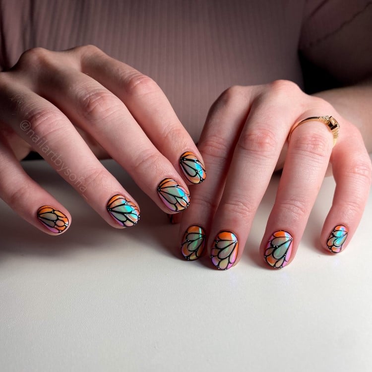 A imagem mostra mãos com unhas decoradas em um estilo que lembra vitrais. As unhas têm desenhos coloridos em tons de azul, laranja e rosa, com contornos pretos, criando um efeito de mosaico. As mãos estão sobre uma superfície clara, e uma delas usa um anel dourado, adicionando um toque de elegância.