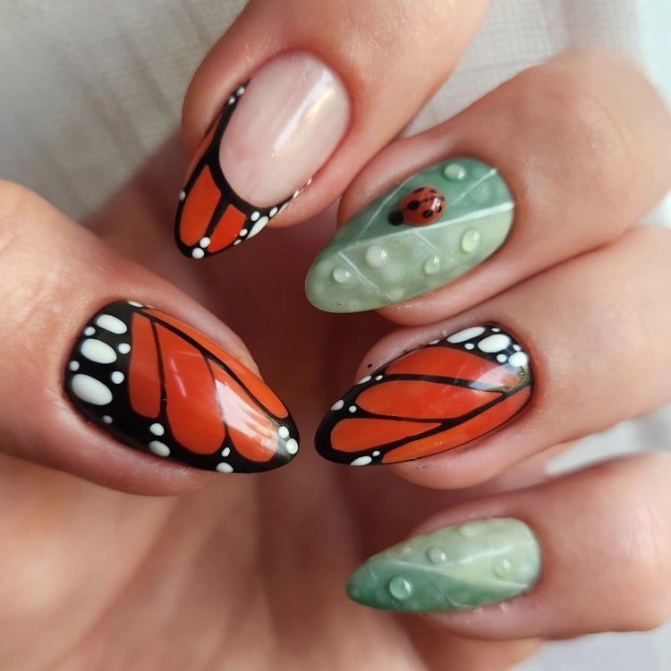 A imagem mostra unhas decoradas com design de asas de borboleta e folhas. Três unhas têm um padrão de borboleta em laranja e preto, com detalhes em branco. As outras duas unhas são pintadas de verde, imitando folhas, com uma pequena joaninha vermelha em uma delas. O visual é detalhado e vibrante.