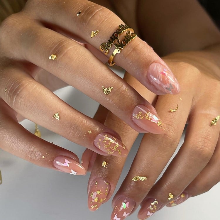 A imagem mostra duas mãos com unhas longas e pontiagudas, decoradas com flocos dourados sobre um esmalte rosa claro. Algumas unhas têm detalhes em branco e rosa que lembram pétalas. As mãos estão adornadas com um anel dourado no dedo médio, com uma inscrição. Flocos dourados também estão espalhados sobre a pele das mãos.