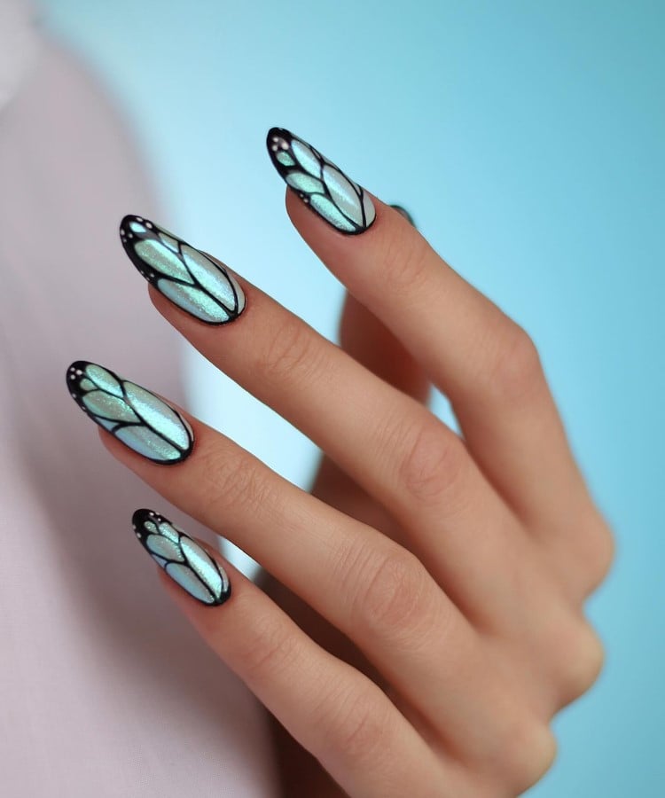 A imagem mostra uma mão com unhas longas decoradas em um estilo que lembra asas de borboleta. As unhas são pintadas em azul claro, com contornos pretos que formam padrões semelhantes a vitrais. Pequenos pontos brancos adornam as pontas, acrescentando detalhes delicados. O fundo é azul claro, destacando o design elegante e artístico das unhas.