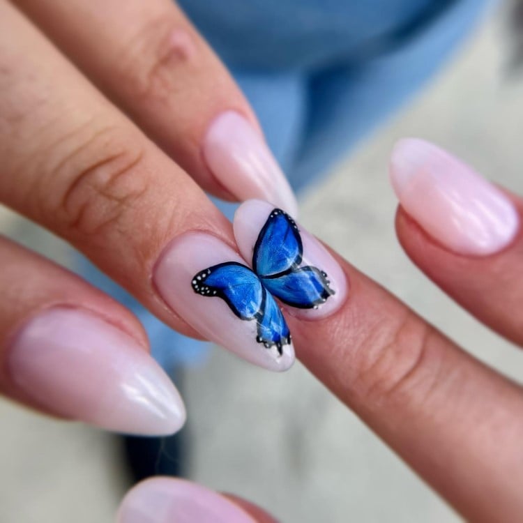 A imagem mostra unhas longas e ovais, pintadas com esmalte rosa claro. No dedo anelar, há uma borboleta azul vibrante desenhada, com detalhes pretos e brancos nas asas. As outras unhas mantêm um tom uniforme e brilhante. O fundo é desfocado, com tons de azul e cinza, destacando o design delicado das unhas.