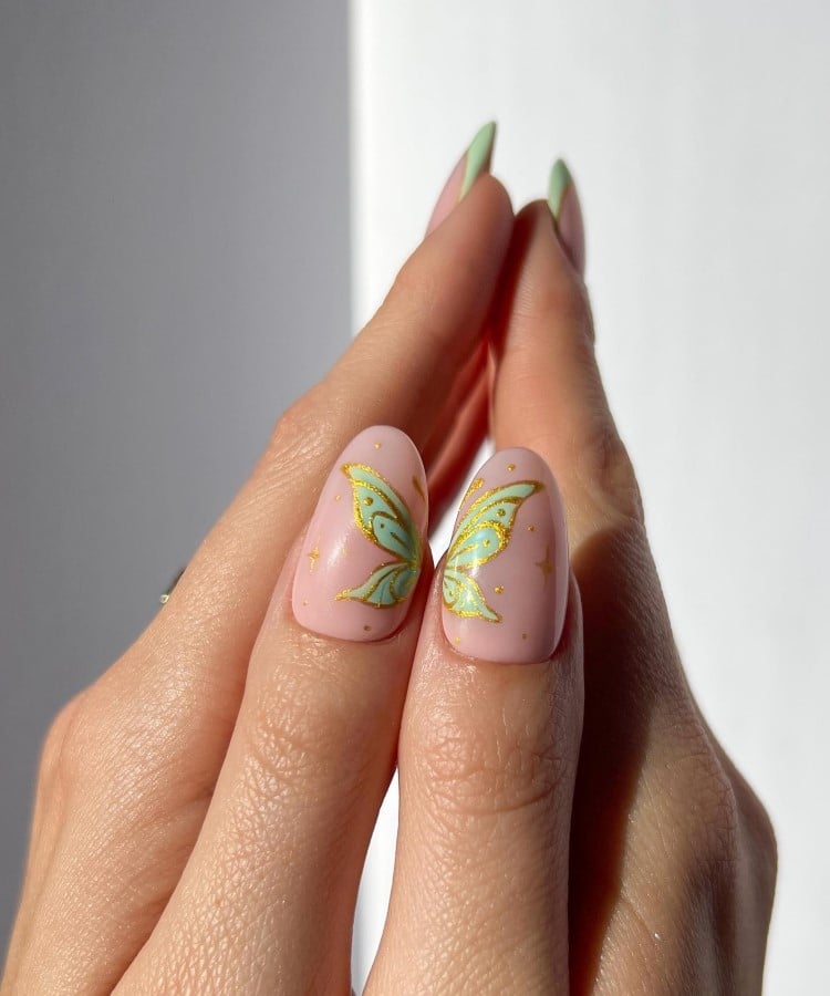 A imagem mostra duas mãos unidas, com unhas longas e ovais. As unhas dos polegares têm um fundo rosa claro e estão decoradas com desenhos de borboletas em verde e amarelo, criando um efeito espelhado quando juntas. As outras unhas são pintadas de verde nas pontas. A iluminação é suave, destacando o design delicado.