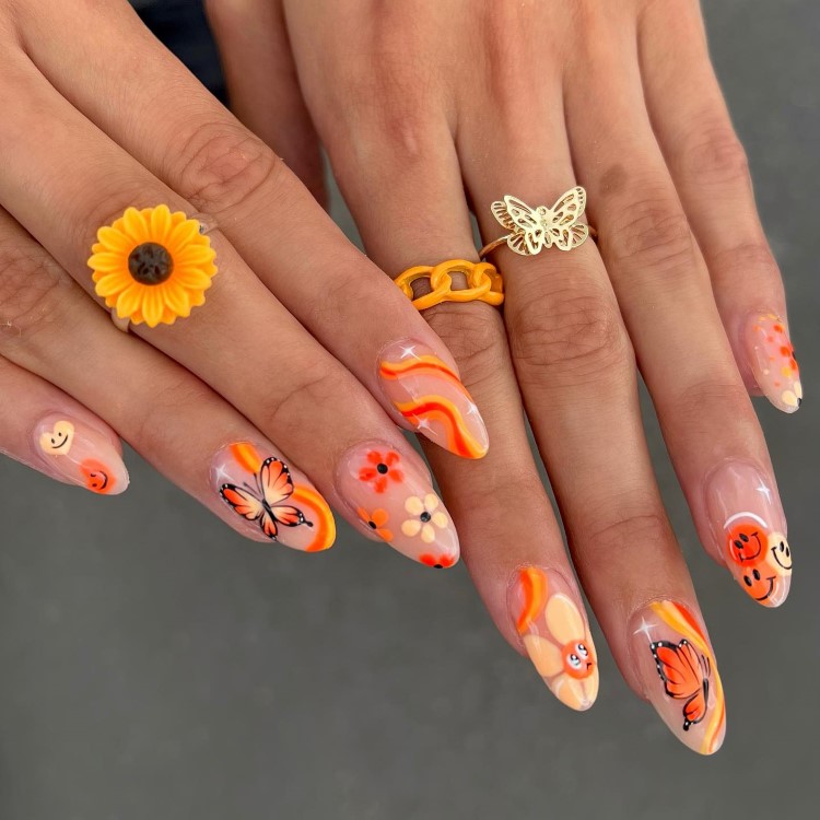 A imagem mostra duas mãos com unhas longas e pontiagudas, decoradas em tons de laranja, amarelo e rosa. As unhas têm desenhos de borboletas, flores e rostos sorridentes. Há anéis nos dedos: um de girassol amarelo, outro de corrente laranja e um com uma borboleta dourada. O fundo é cinza, destacando as cores vibrantes.