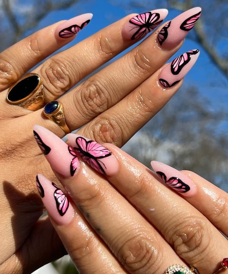 A imagem mostra mãos com unhas longas e pontiagudas, decoradas com desenhos de borboletas em tons de rosa e preto. As unhas têm um fundo rosa claro, destacando as borboletas estilizadas. As mãos exibem anéis dourados com pedras pretas e azuis. O fundo é um céu azul claro, adicionando contraste à cena.