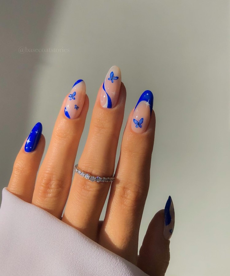 A imagem mostra uma mão com unhas longas e ovais. As unhas têm um design moderno com esmalte azul vibrante nas pontas e desenhos de pequenas borboletas azuis sobre um fundo nude. O dedo anelar exibe um anel prateado com pedras brilhantes. A manga da roupa é branca, e o fundo é suave e claro.