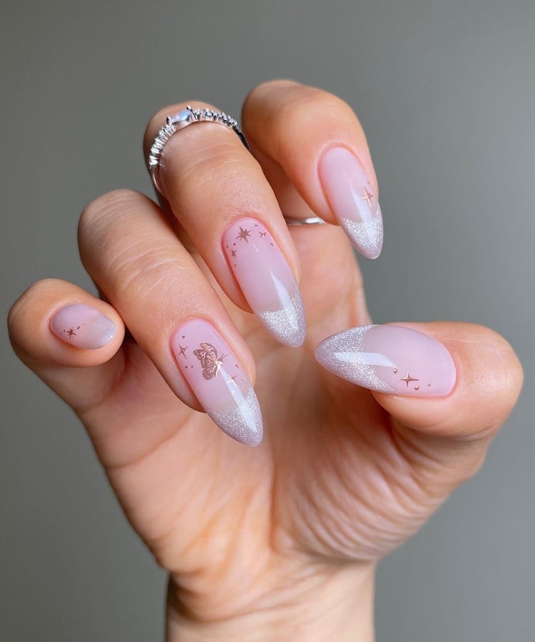 A imagem mostra uma mão com unhas longas e em formato amendoado, decoradas com esmalte rosa claro e pontas prateadas cintilantes. Pequenos desenhos de estrelas e uma borboleta adornam algumas unhas. A mão usa anéis prateados delicados nos dedos médio e anelar. O fundo é neutro, destacando o elegante design das unhas.