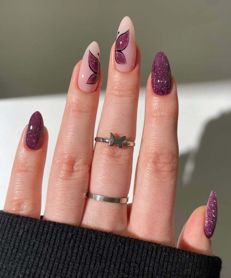 A imagem mostra uma mão com unhas longas e pontiagudas. As unhas do polegar, indicador e mindinho são pintadas de roxo com brilho. As unhas do dedo médio e anelar têm um design de asas de borboleta em tons de rosa e preto. A mão usa dois anéis prateados, um com formato de borboleta. O fundo é claro.