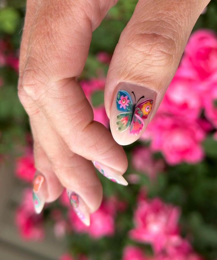 A imagem mostra uma mão com unhas longas e ovais, decoradas com borboletas coloridas. A unha do polegar exibe uma borboleta com asas em tons de azul, rosa e verde, com detalhes florais. O fundo é desfocado, cheio de flores rosa vibrantes, destacando o design artístico e delicado das unhas.