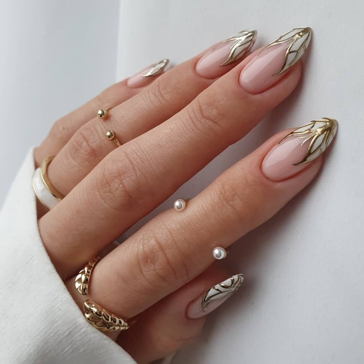 A imagem mostra uma mão com unhas longas e pontiagudas, decoradas em estilo elegante. As unhas têm um esmalte nude com detalhes dourados e brancos em formato de asas nas pontas. A mão usa vários anéis dourados finos, alguns com pérolas. A manga da roupa é branca, e o fundo é claro, destacando a sofisticação do visual.