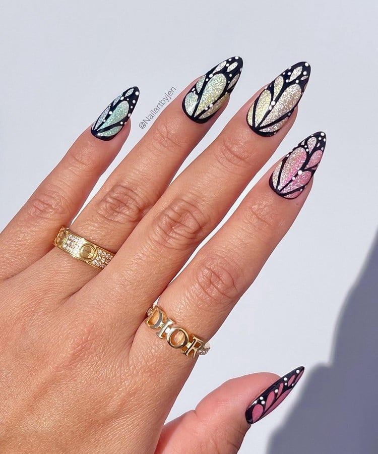 A imagem mostra uma mão com unhas longas e pontiagudas, decoradas em um estilo de asas de borboleta. As unhas têm um fundo brilhante em tons de azul, verde, amarelo e rosa, com contornos pretos e pequenos pontos brancos. A mão usa dois anéis dourados, um deles com a inscrição "DIOR". O fundo é branco.