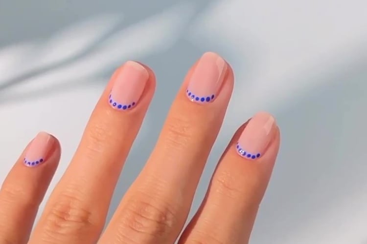 A imagem mostra uma mão com unhas decoradas. As unhas têm um esmalte base em tom claro e estão adornadas com pequenas bolinhas azuis formando uma linha curva próxima à ponta. O fundo é suave e neutro, destacando o design delicado e minimalista das unhas.